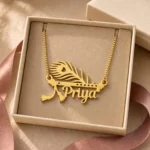 Gold-tone peacock feather and flute name pendant displayed in an open beige jewellery box