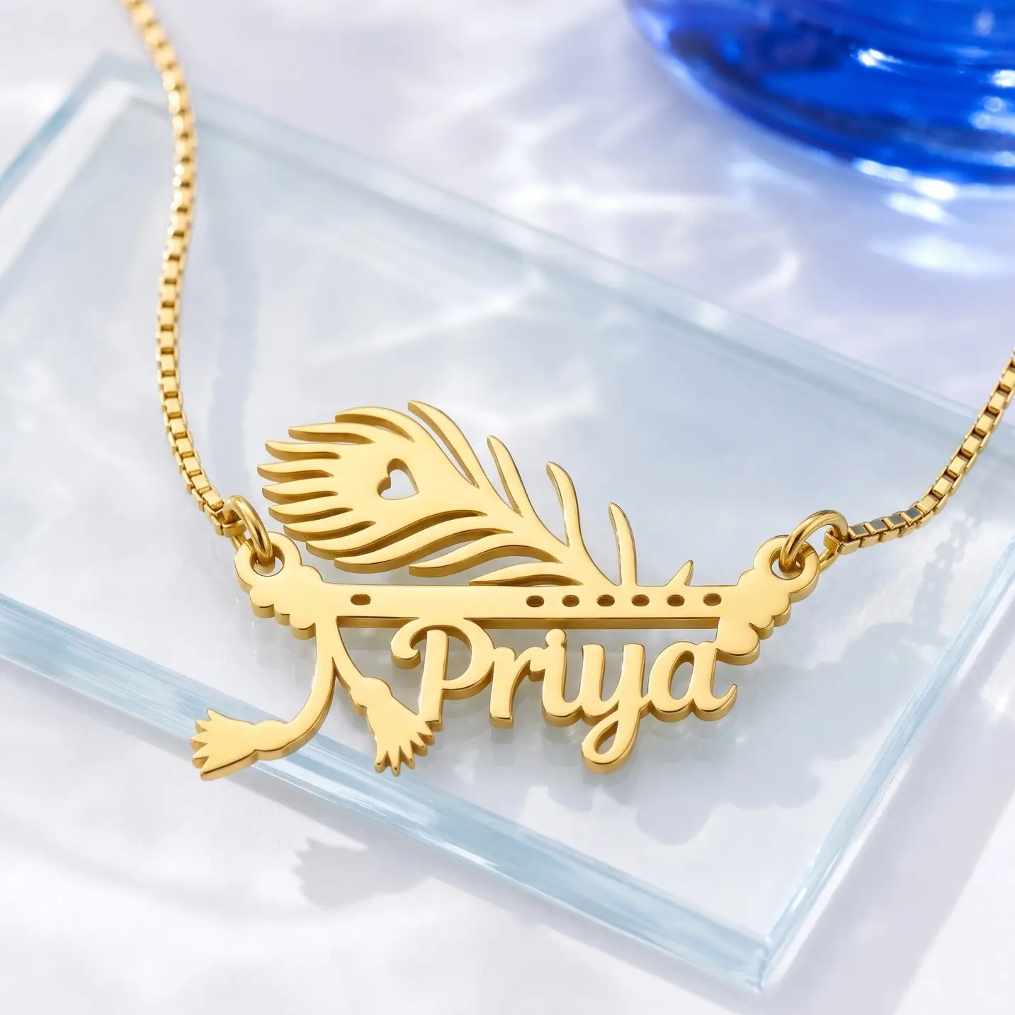 Mor Pankh Personalized Name Pendant - Glass Display Gold-tone peacock feather and flute name pendant on a clear glass display