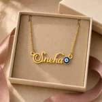 Gold-tone name necklace with a blue evil eye accent displayed in an open beige jewellery box