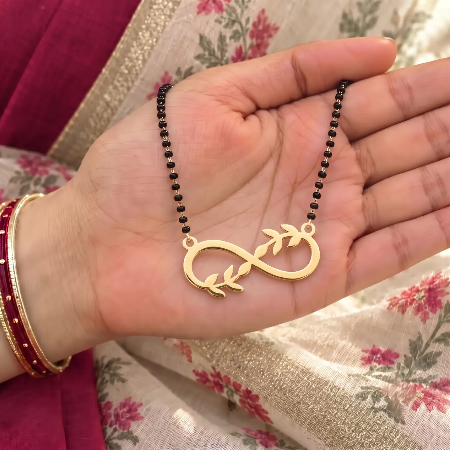 Leaf Design Infinity Necklace - Handheld View Gold-tone infinity pendant with leaf accents on a black-bead chain held in a hand