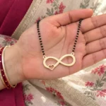 Gold-tone infinity pendant with a small side heart on a black-bead chain held in a hand