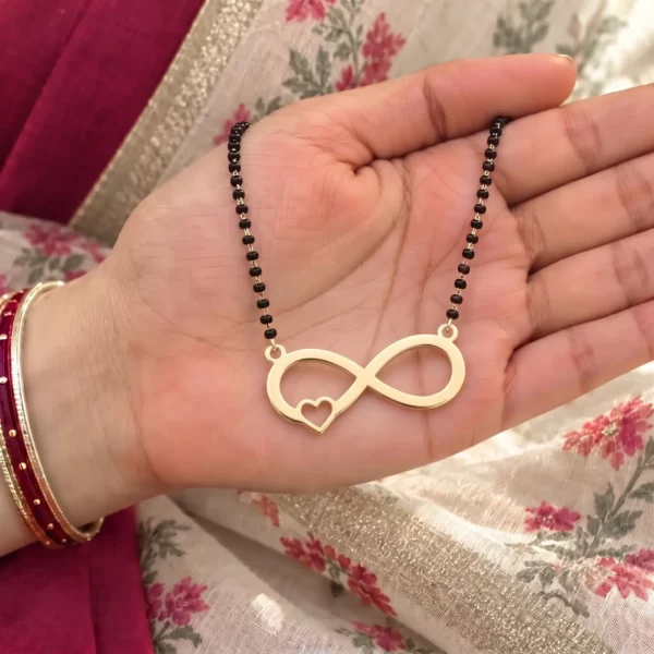 Gold-tone infinity pendant with a small side heart on a black-bead chain held in a hand