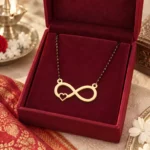 Gold-tone infinity pendant with a small side heart on a black-bead chain displayed in a red jewellery box