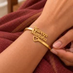Open Heart Couple Name Bracelet in a golden finish, worn on a wrist beside maroon fabric