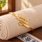 Open Heart Couple Name Bracelet in a golden finish, displayed on rolled cream fabric