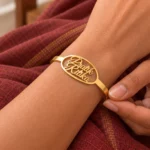 Gold-tone couple name bracelet with two script names inside an oval frame, worn on a wrist beside maroon fabric