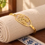 Gold-tone couple name bracelet with two script names inside an oval frame, displayed on rolled cream fabric